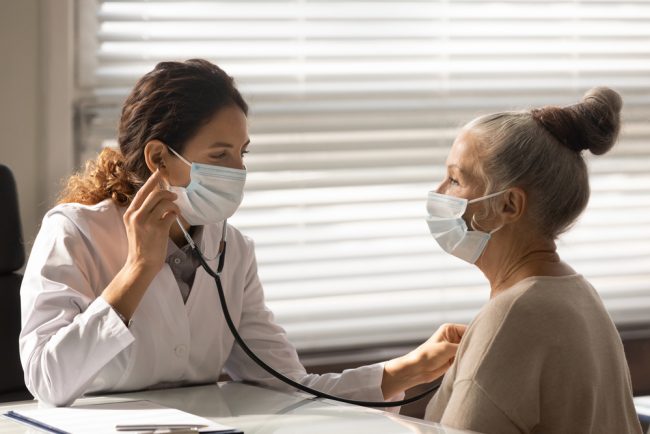 Caring,Young,Female,Doctor,In,Medical,Facemask,Hold,Stethoscope,Listen